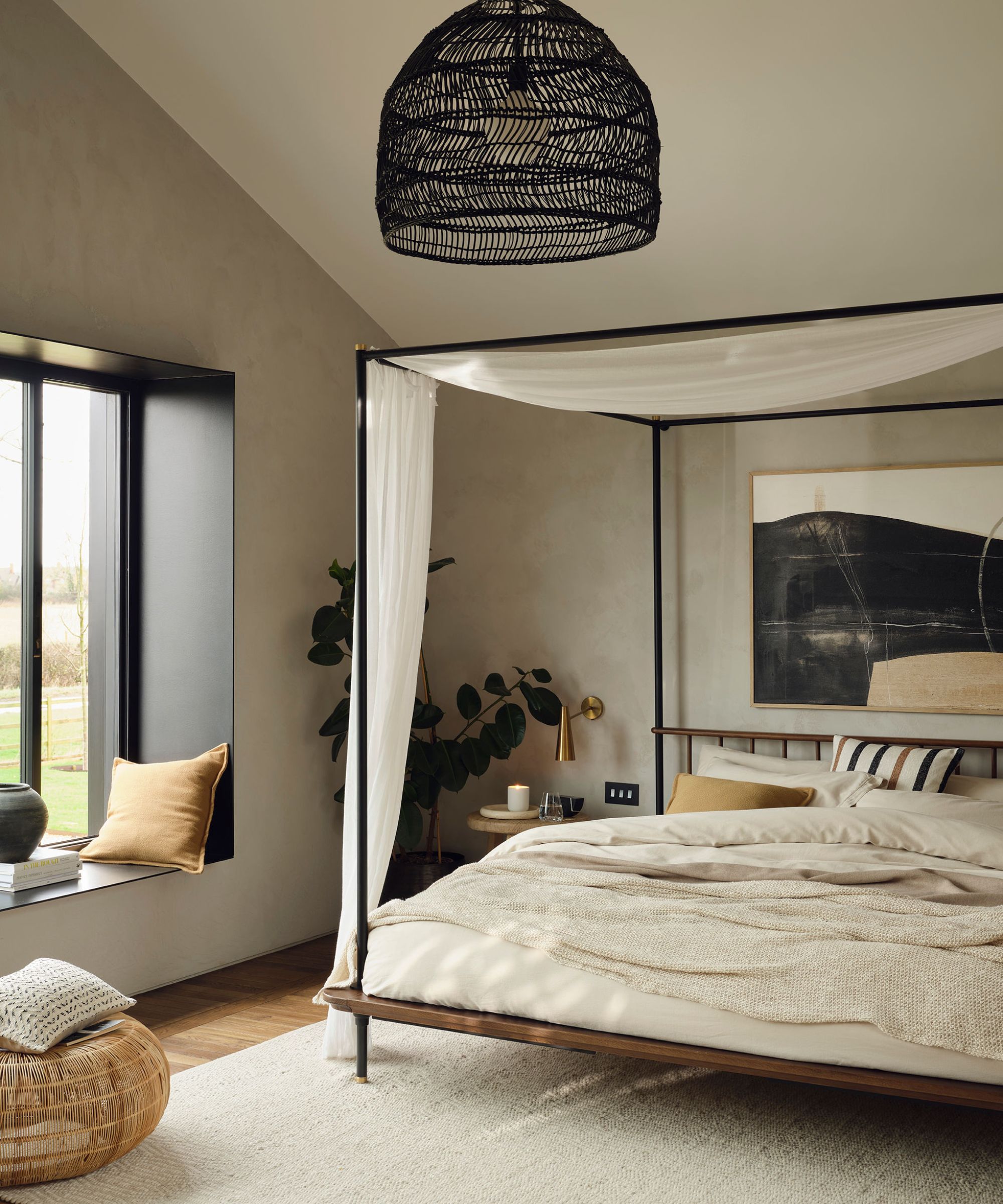 A neutral bedroom with a black four poster bed, a large window with a bench seat
