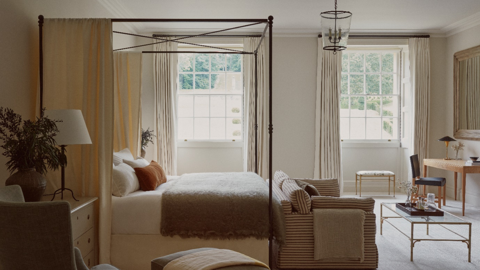 A neutral bedroom with a thin wooden four-poster bed, a couch at the foot, and a small glass coffee table. 