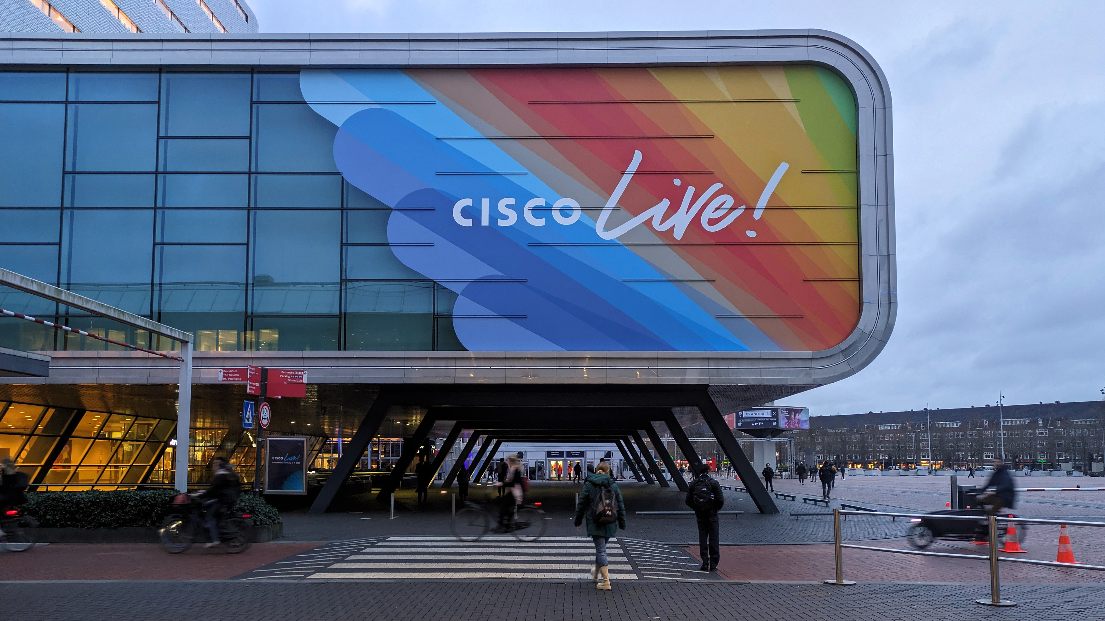 The Cisco Live 2024 sign on the outside of the RAI conference center in Amsterdam, against a background of rainbow colors. Below, cyclists move through the street.