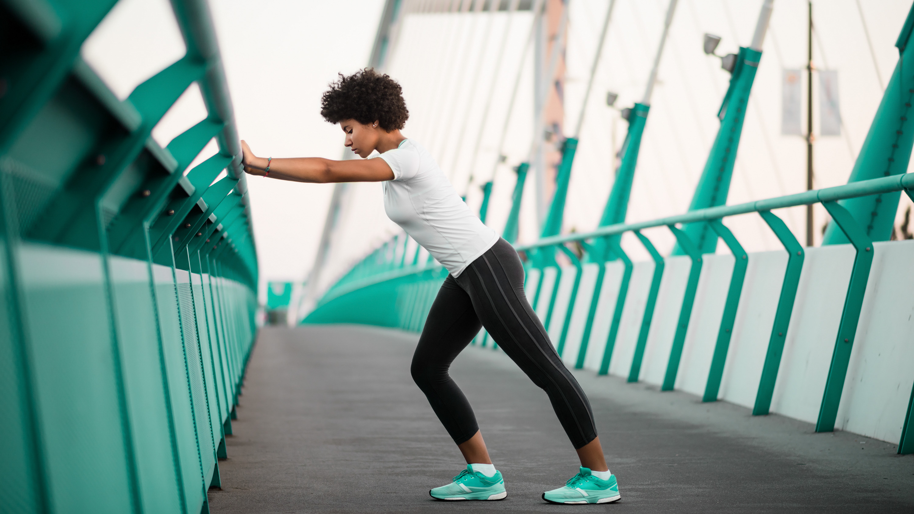 A woman stands doing a warm-up on a bridge. She is leaning against the bridge with her hands, with her left leg extended behind her and her right leg gently bent in front. 