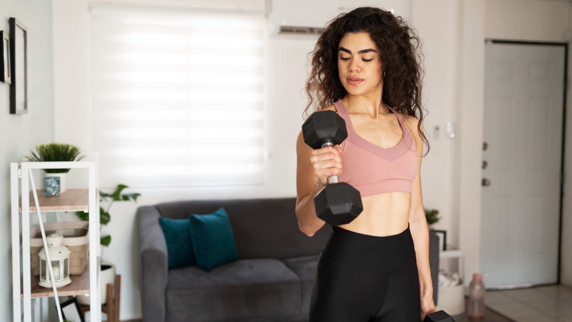 woman curling a dumbbell up to her biceps standing in a living room setting. she&#039;s wearing a light pink crop top and dark bottoms. 