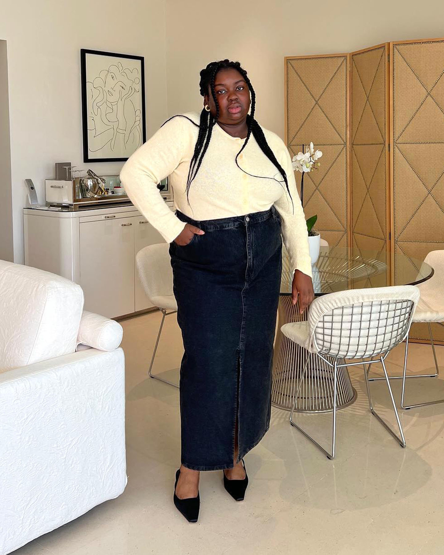 British female fashion influencer and creative director Abisola Omole poses in a modern London apartment wearing hoop earrings, a pale yellow buttoned cardigan, dark denim maxi skirt, and black kitten heel slingbacks