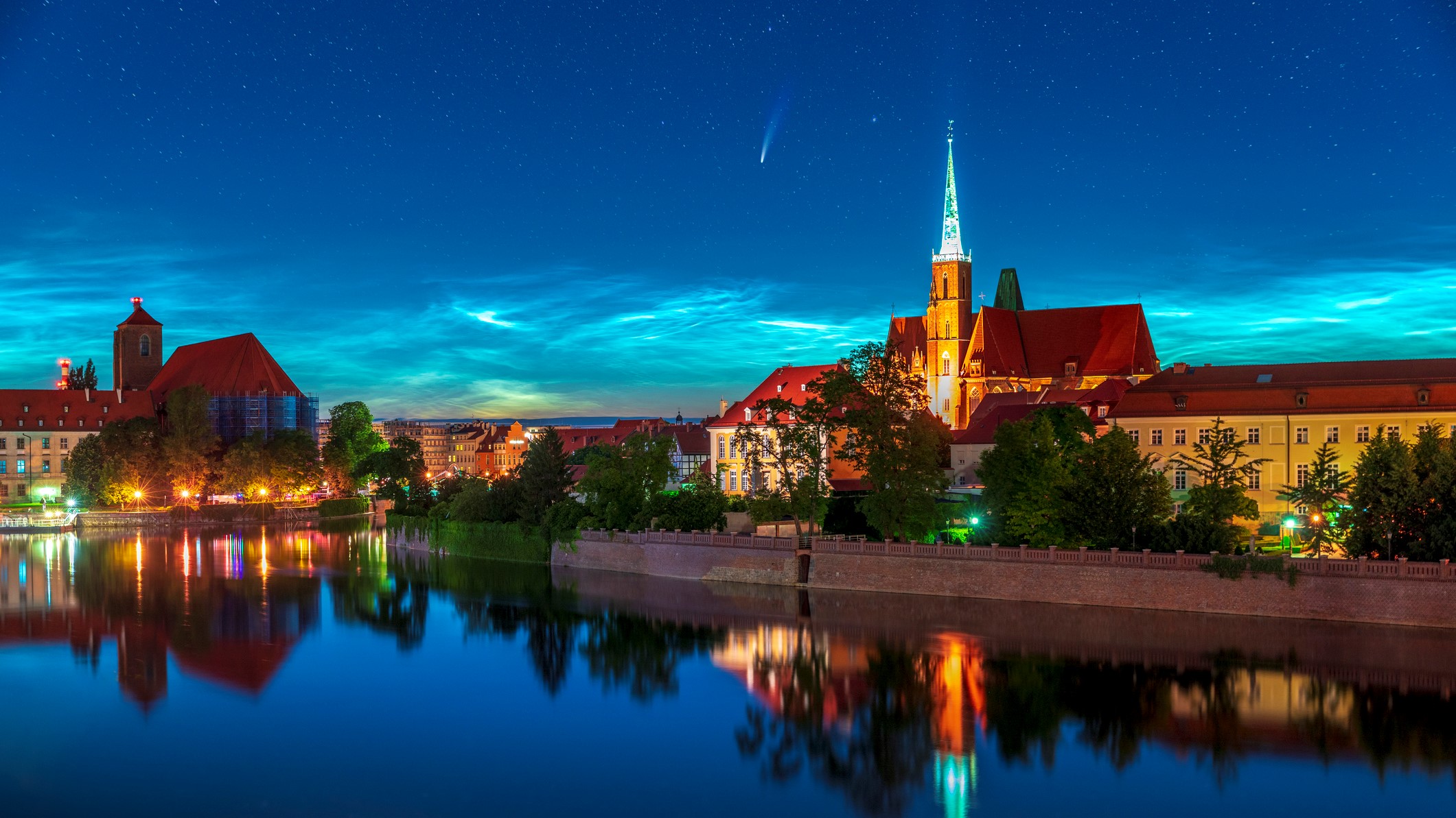 Noctilucent clouds are visible at astronomical twilight. The image shows light wispy clouds in dark blue sky with comet NEOWISE flying overhead with a long white tail.