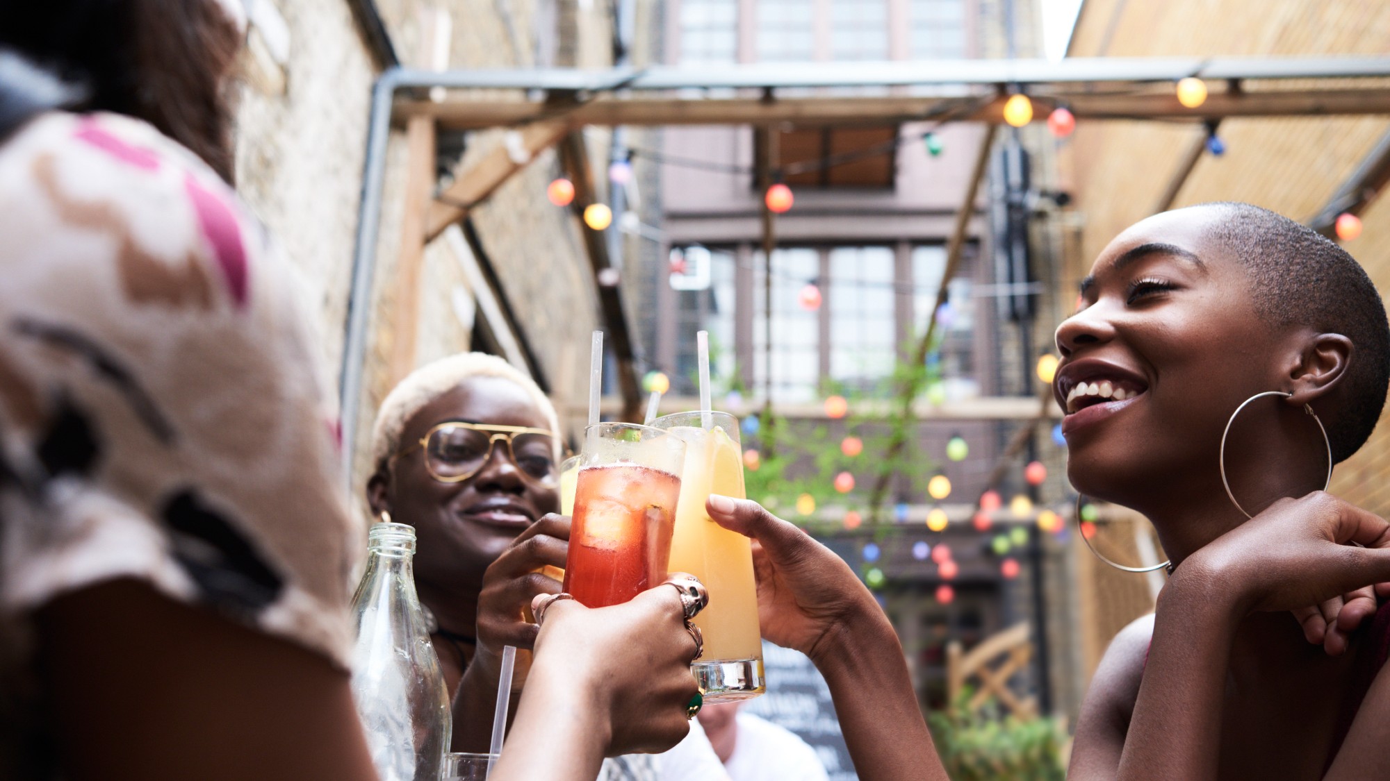 three Black women cheersing with highball glasses filled with cocktails in an outdoor setting