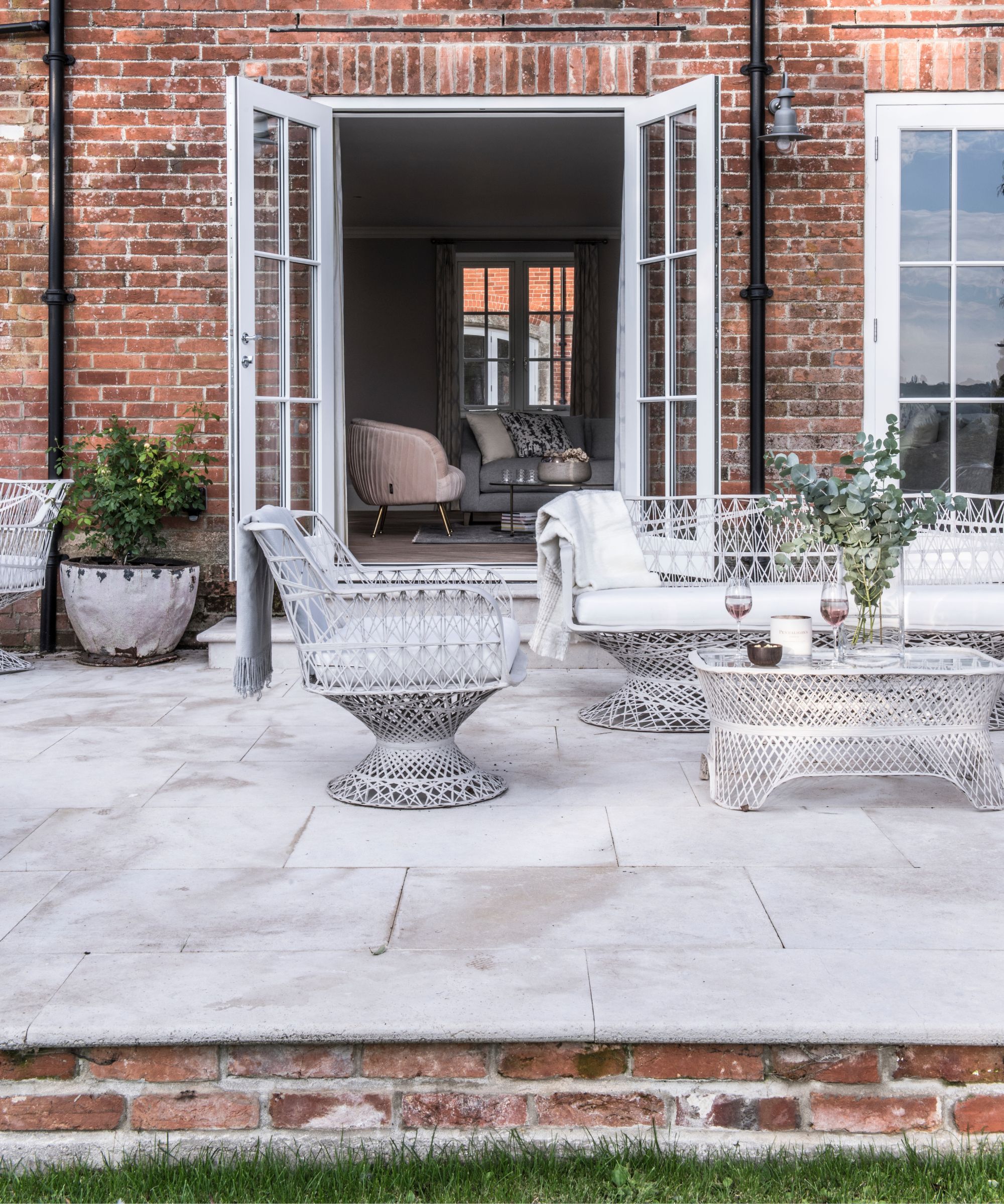 A raised paved patio outside a red brick house with white rattan furniture and open french doors with a view into a bright and modern living room