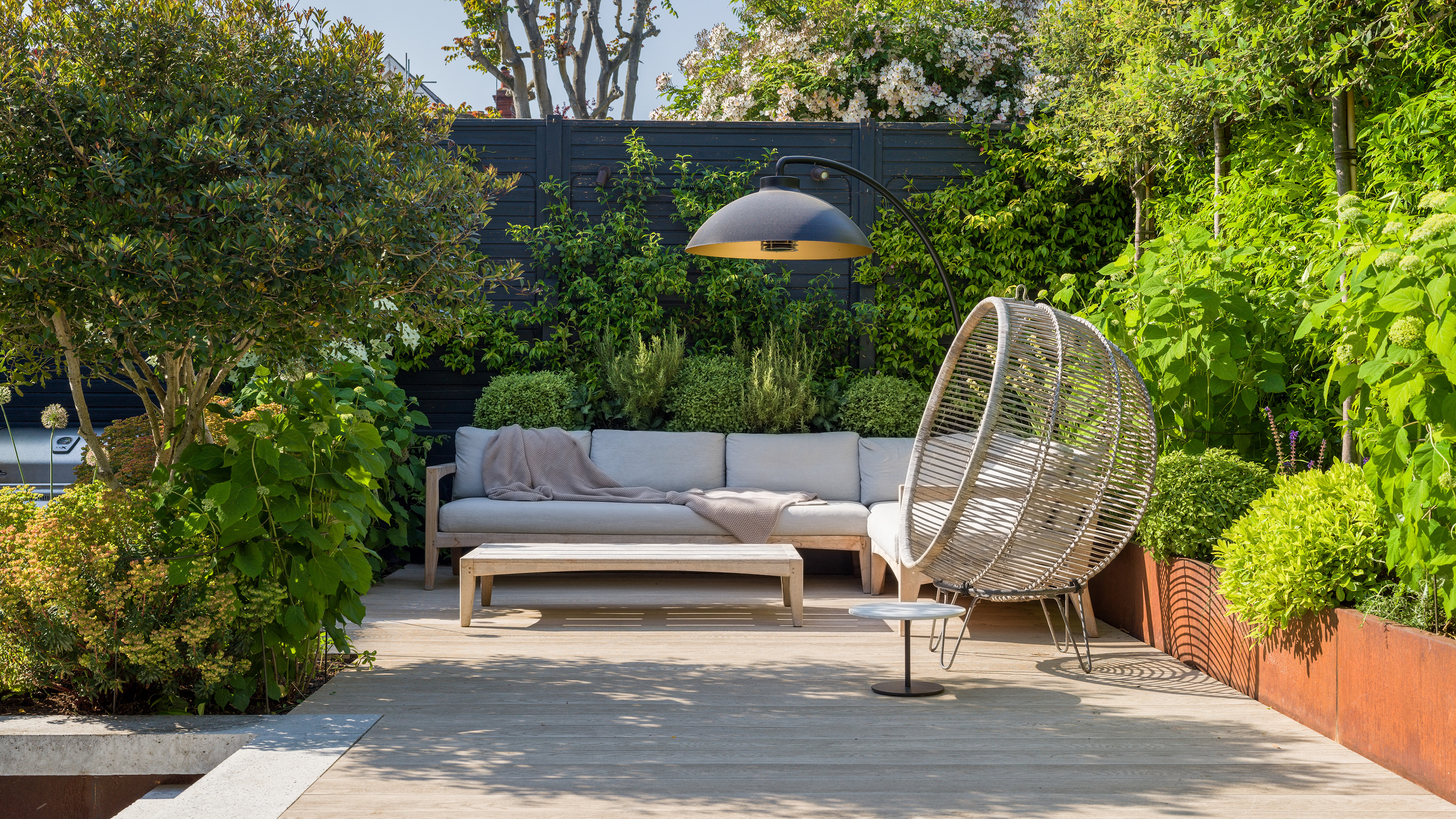 white outdoor sofa with coffee table, egg chair and corten steel planters surrounded by pleached trees and a black fence