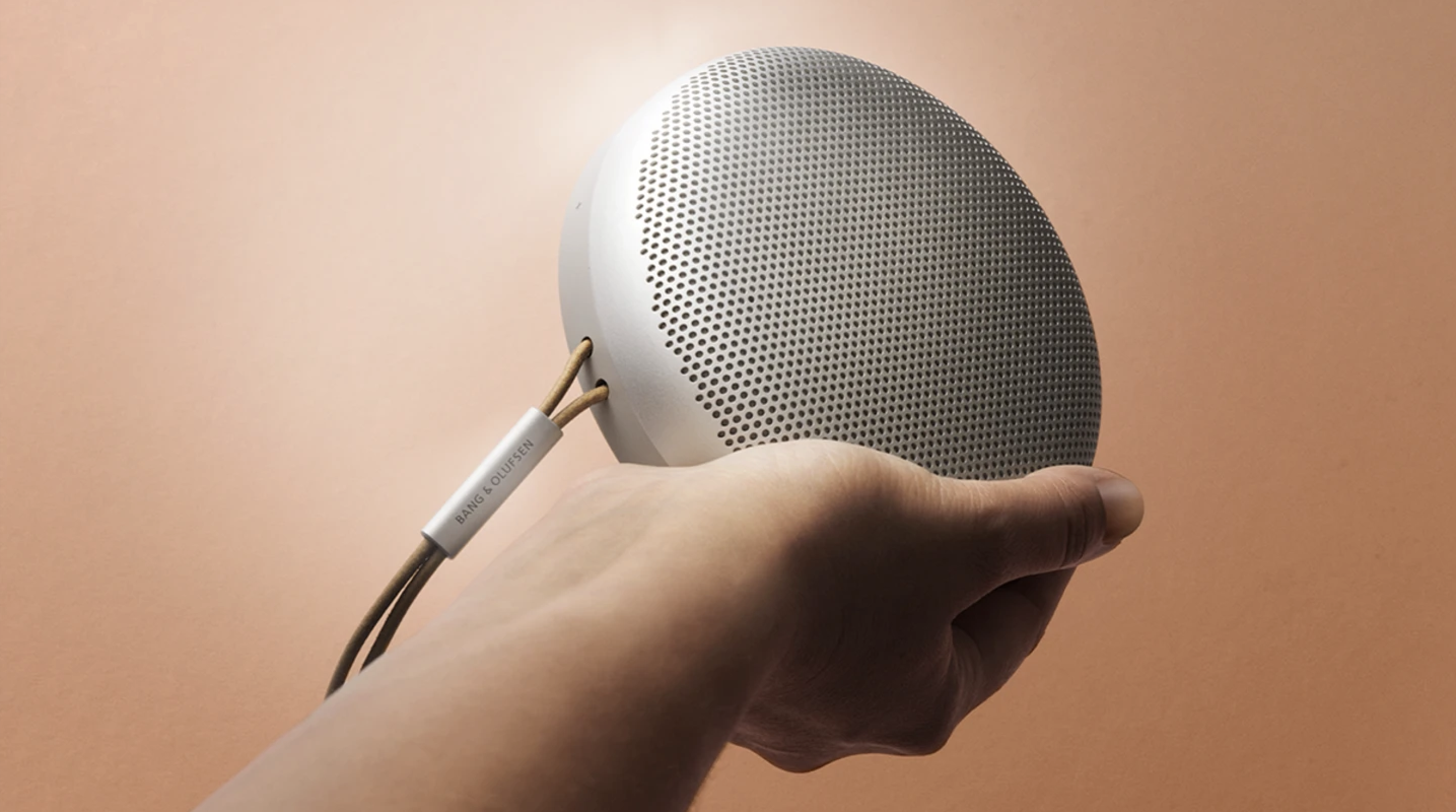 A hand holding a silver Bang & Olufsen Beosound A1 (2nd Gen) speaker. It is round with a strap attached. The background is light brown.
