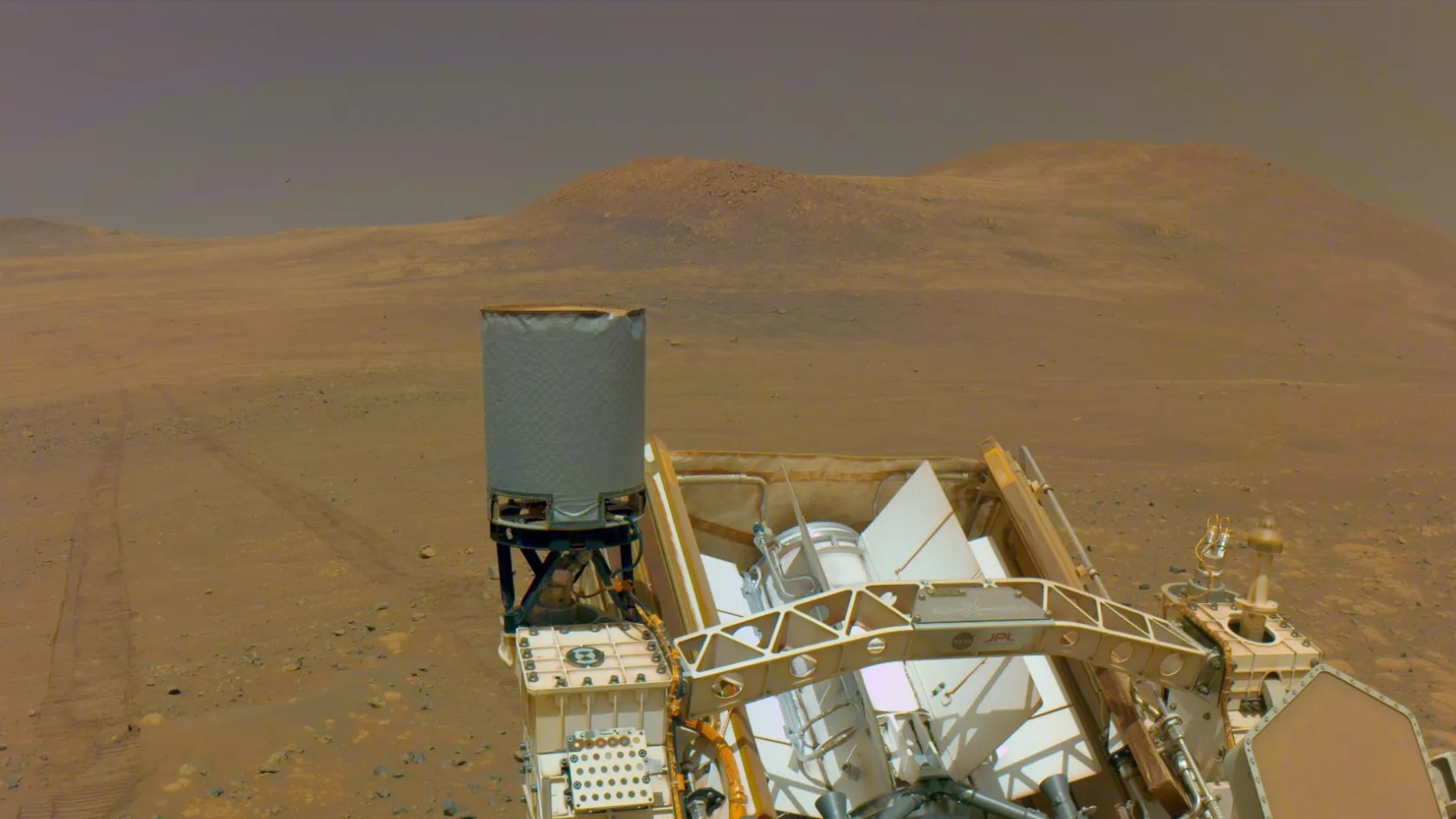 selfie taken by a mars rover, showing bits of its hardware in the foreground and rover tracks extending across a barren reddish-sand landscape in the background