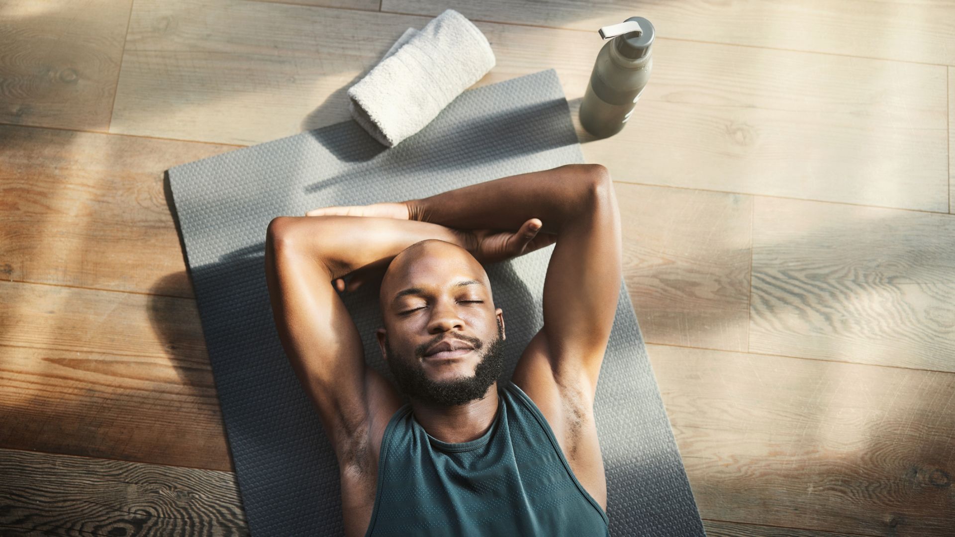 man lies on yoga mat with arms folded above head and with eyes closed