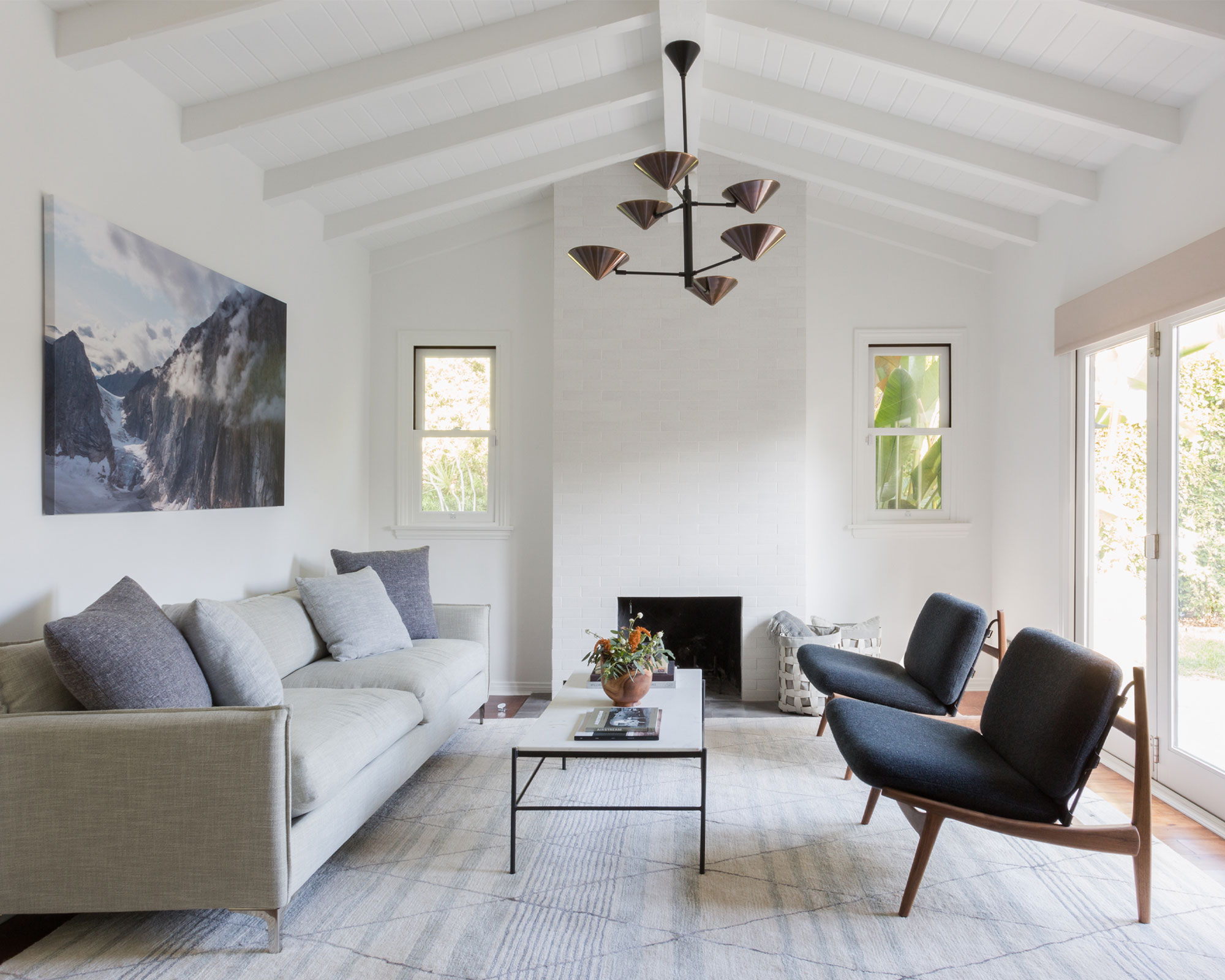 White painted living room with white wooden paneling and beams, large striped rug, cream sofa and twin lounge chairs, white and black coffee table, metallic hanging pendant, mountainscape artwork