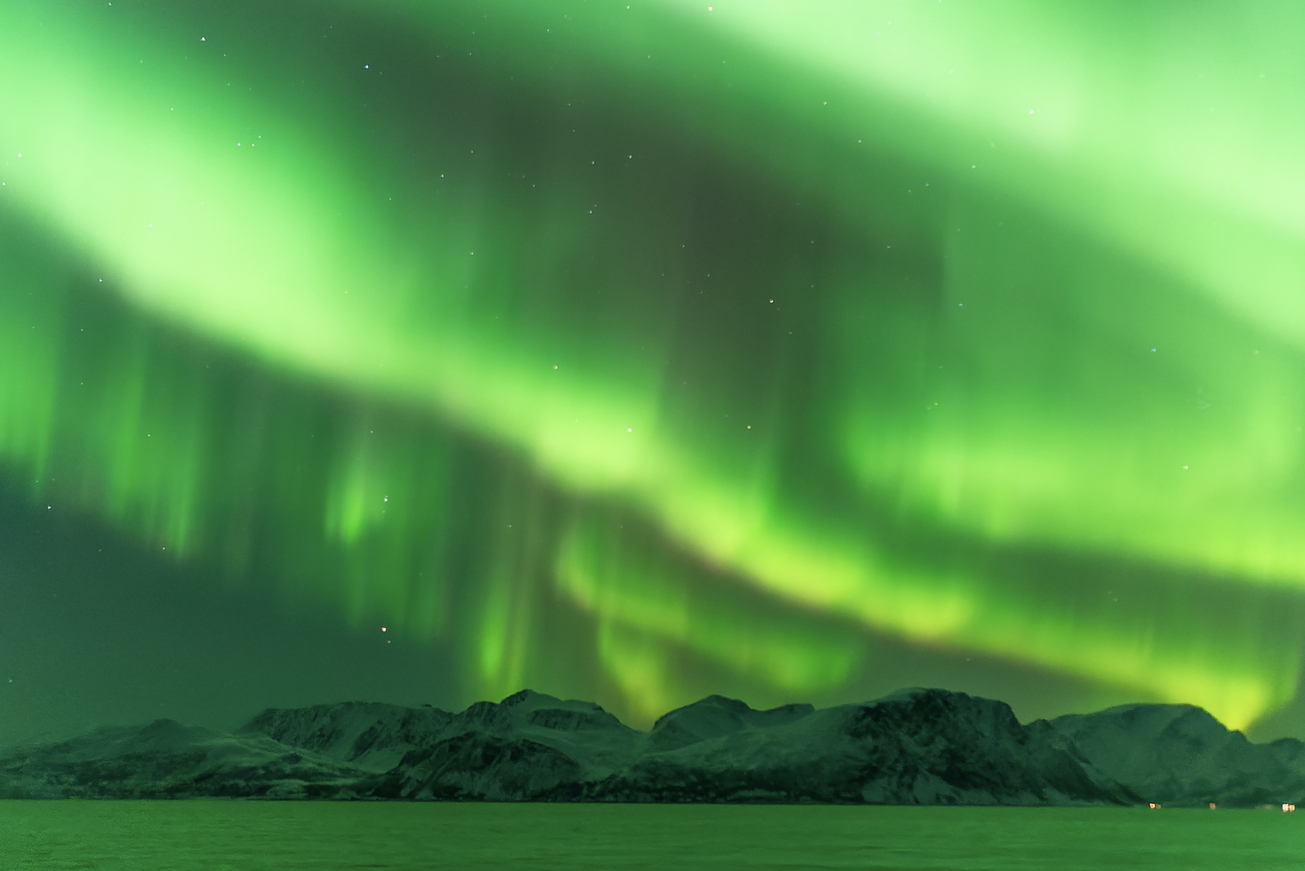 northern lights in Norway appearing as vivid green bands of light across the sky. With mountains and water below.