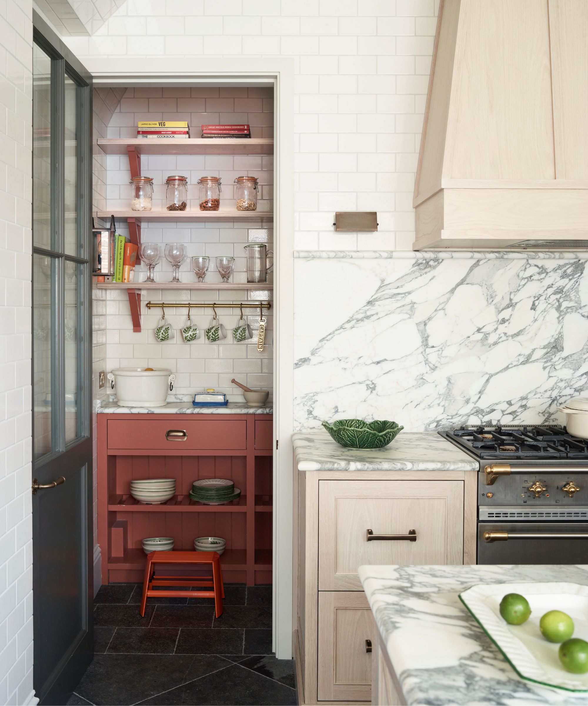 A marble kitchen with island, baby pink drawers and range hood, white tiles, and a grey hob. To the left hand-side you can see a tiled white pantry with pale red cabinets, and shelves with glasses and recipe books.