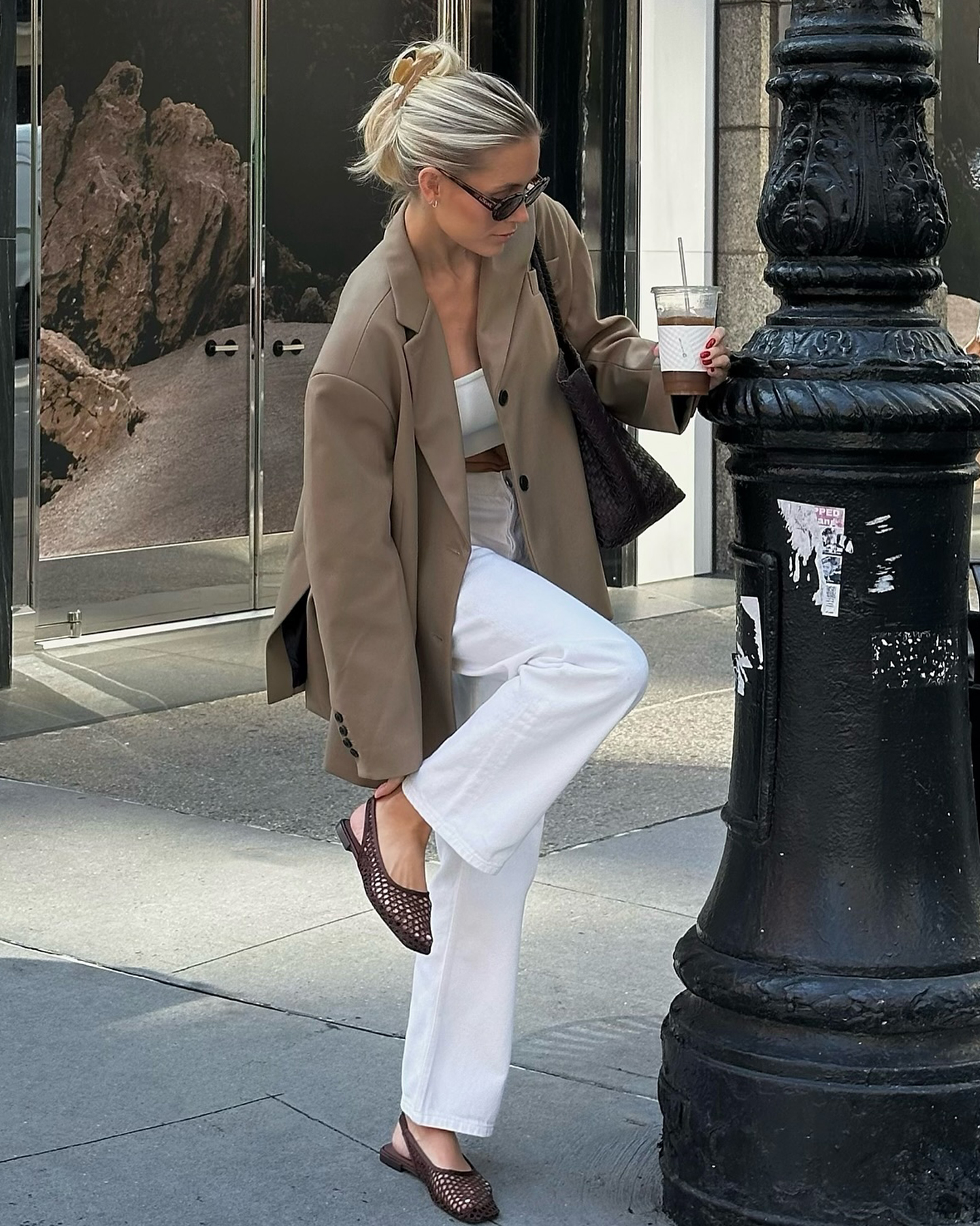 influencer Fredrika Ekerot poses on a NYC sidewalk wearing a hair claw clip, sunglasses, light brown blazer, white crop top, white straight-leg jeans, and brown woven slingback flats