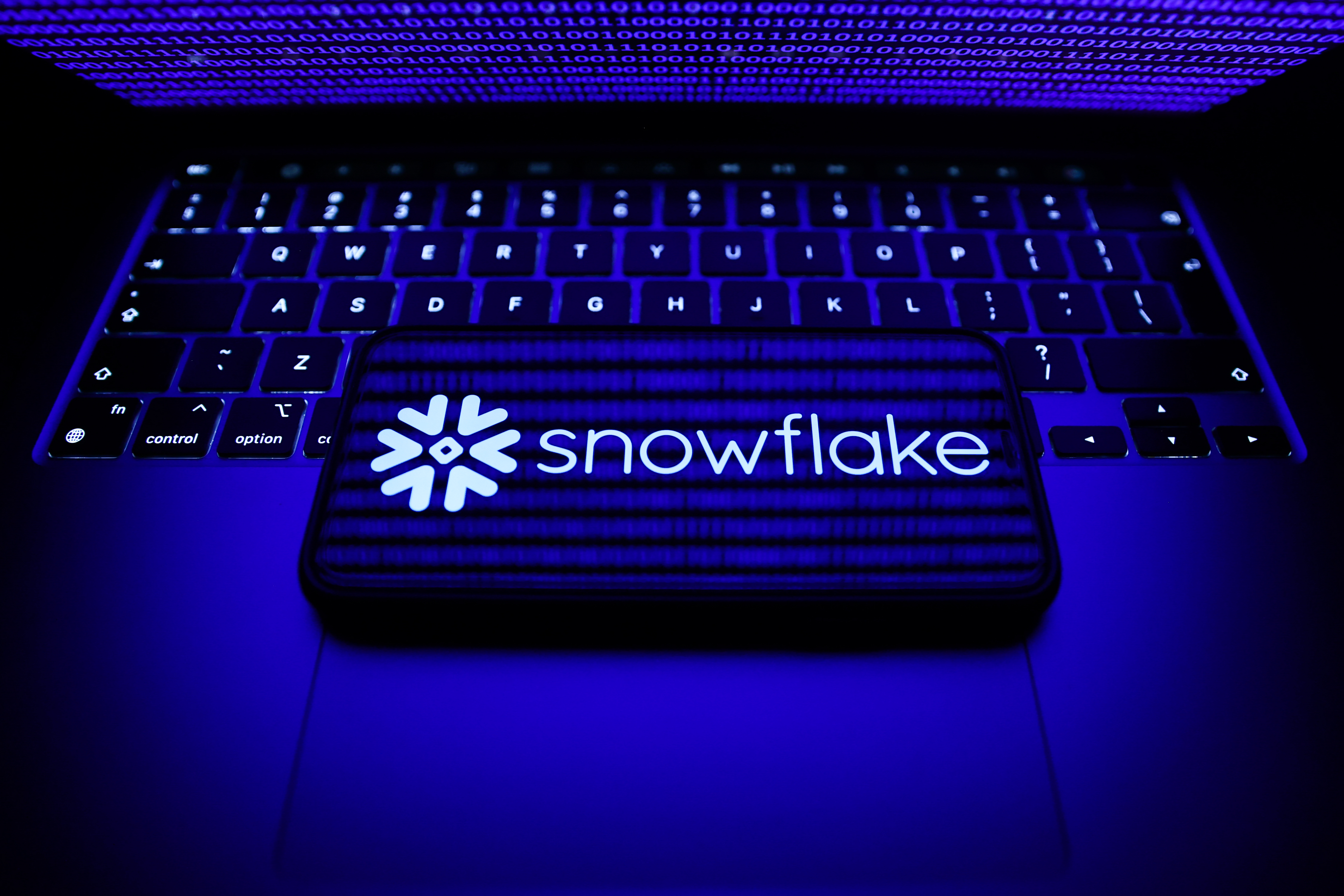 Snowflake company logo appearing on a smartphon that's laid in landscape orientation on top of a MacBook's keyboard with the computer's lid half closed, beaming a blue light onto the keyboard and phone