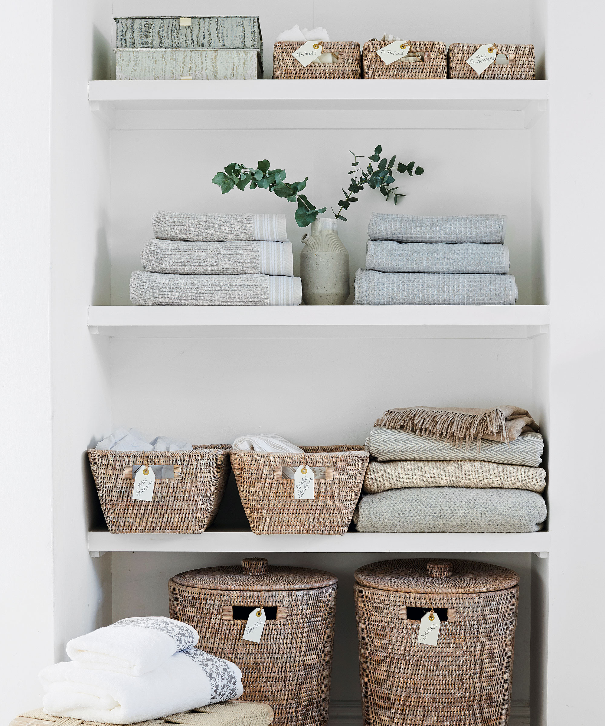White alcove open shelving with neatly folder piles of towels, linens, blankets and wicker storage baskets with labels on